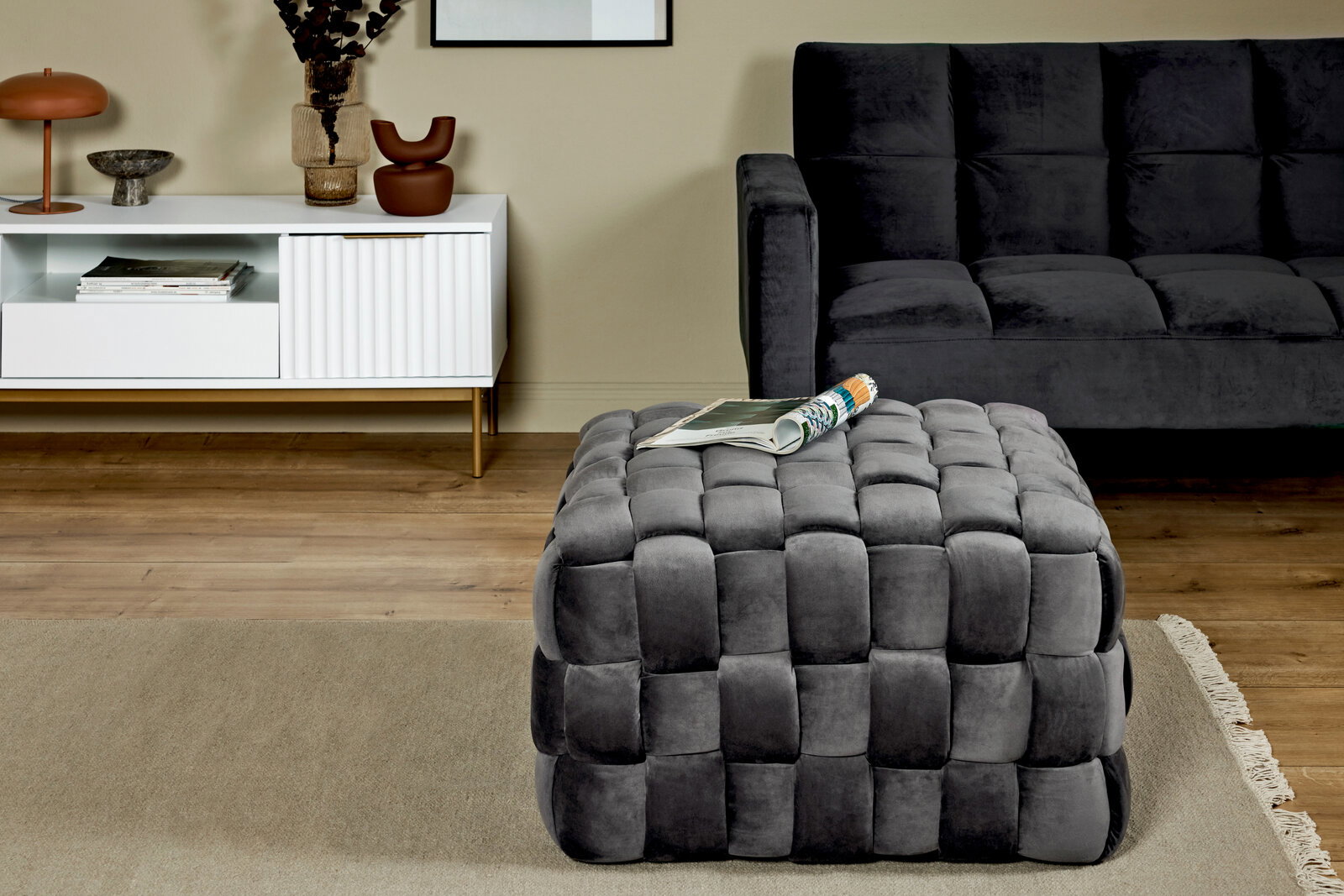 Grauer, gepolsterter Hocker in Würfelform mit geflochtenem Design, im Vordergrund eines modernen Wohnzimmers mit seitlicher Perspektive.