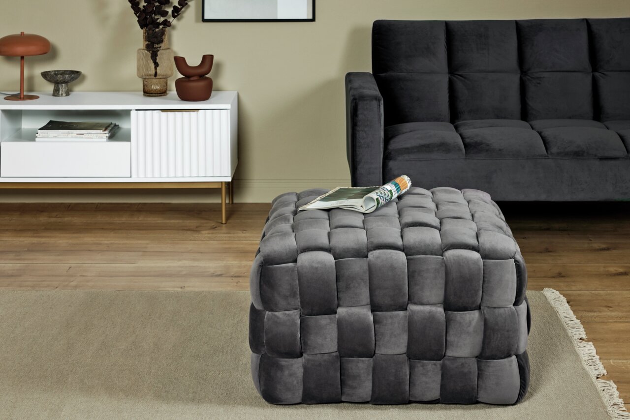 Grijze, gestoffeerde kubusvormige poef met gevlochten patroon, op de voorgrond van een moderne woonkamer, gezien vanuit een zijdelingse hoek.