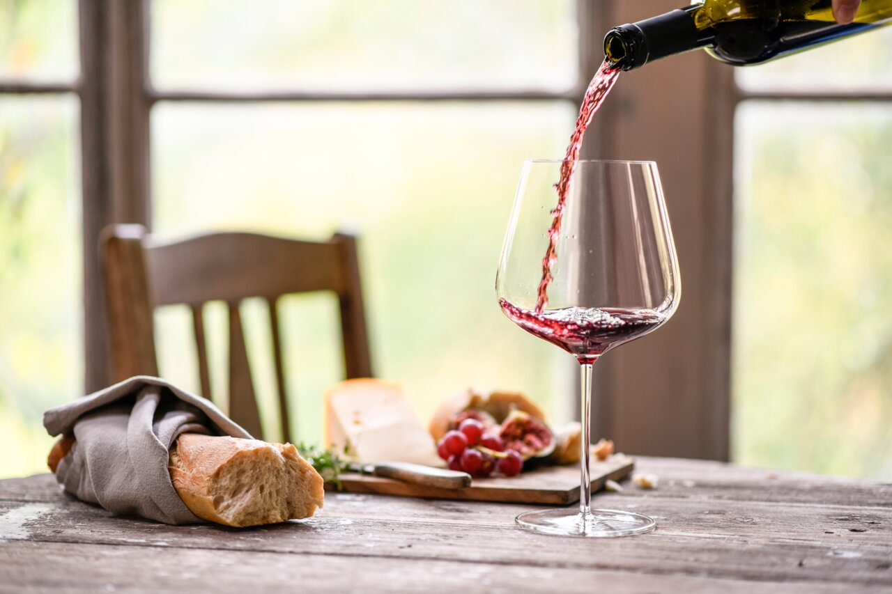 Ein elegantes Burgunder Rotweinglas Vervino, aus dem Rotwein eingeschenkt wird, auf einem rustikalen Holztisch mit Baguette, Käse und Trauben im Hintergrund. Seitliche Perspektive.