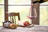 Ein elegantes Burgunder Rotweinglas Vervino, aus dem Rotwein eingeschenkt wird, auf einem rustikalen Holztisch mit Baguette, Käse und Trauben im Hintergrund. Seitliche Perspektive.
