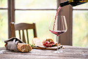 Ein elegantes Burgunder Rotweinglas Vervino, aus dem Rotwein eingeschenkt wird, auf einem rustikalen Holztisch mit Baguette, Käse und Trauben im Hintergrund. Seitliche Perspektive.
