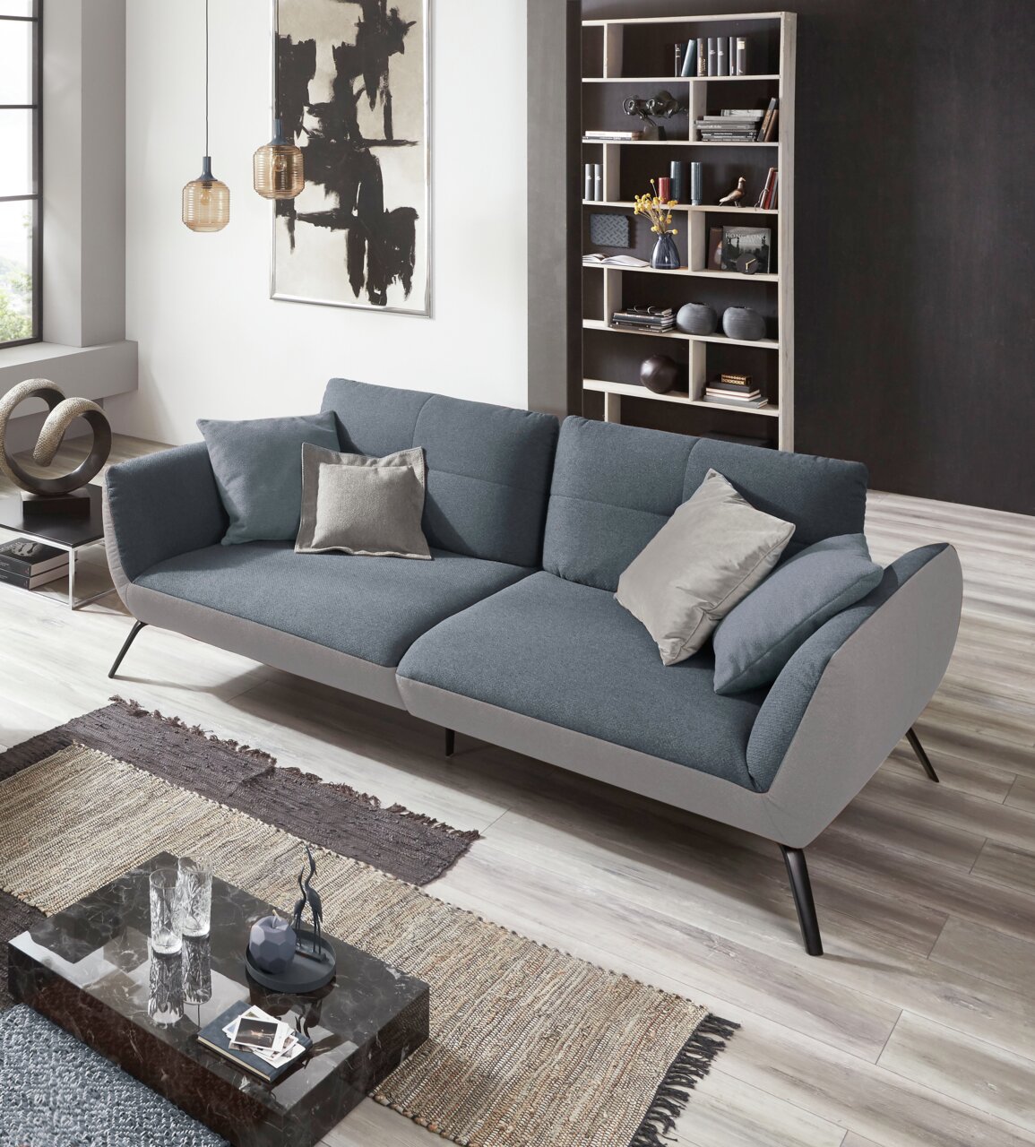 Modernes Sofa in Grau mit Kissen, seitliche Perspektive, in stilvollem Wohnzimmer mit Kunstwerk und Bücherregal.