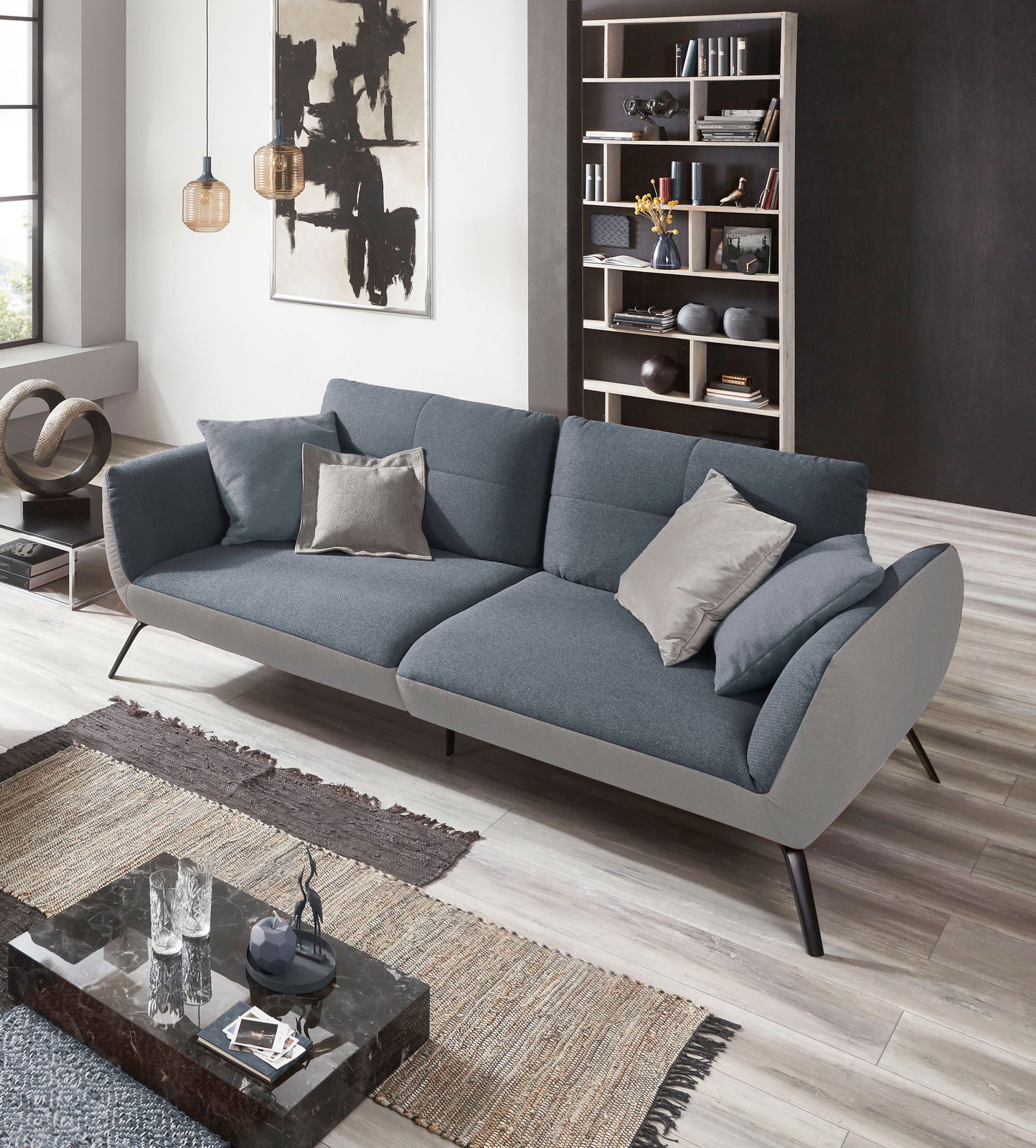 Modernes Sofa in Grau mit Kissen, seitliche Perspektive, in stilvollem Wohnzimmer mit Kunstwerk und Bücherregal.