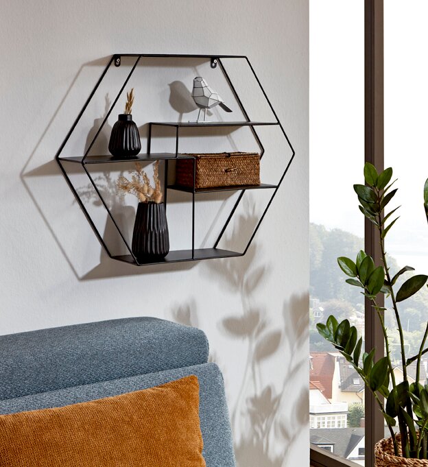 Sechseckiges Wandregal aus schwarzem Metall mit mehreren Ablagen, dekoriert mit zwei schwarzen Vasen, einem kleinen Vogel und einem Korb, an heller Wand über Sofa frontale Ansicht mit leichter Seitenperspektive.