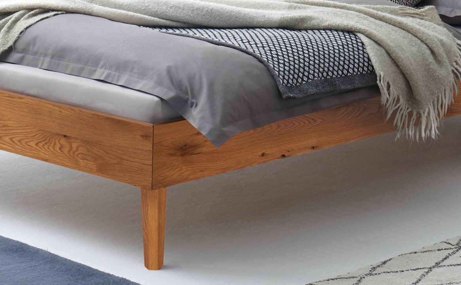 Nahaufnahme eines Bettgestells aus Holz mit Bettwäsche in Grau und einer Decke, seitliche Perspektive