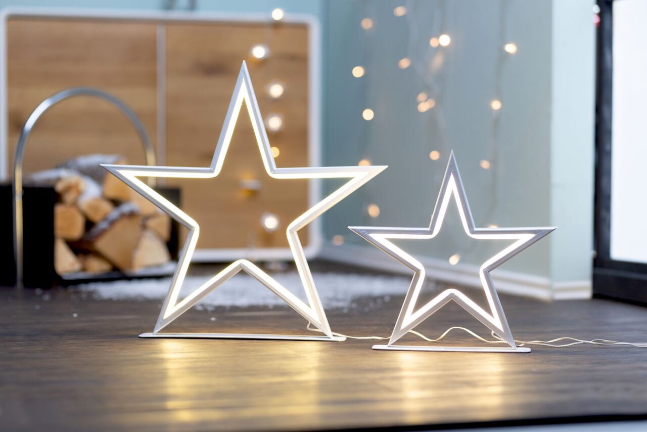 Twee stralende witte LED-sterrenlampjes op de vloer, voor een stapel haardhout en een lichtslinger, van voren gefotografeerd vanuit een laag standpunt.