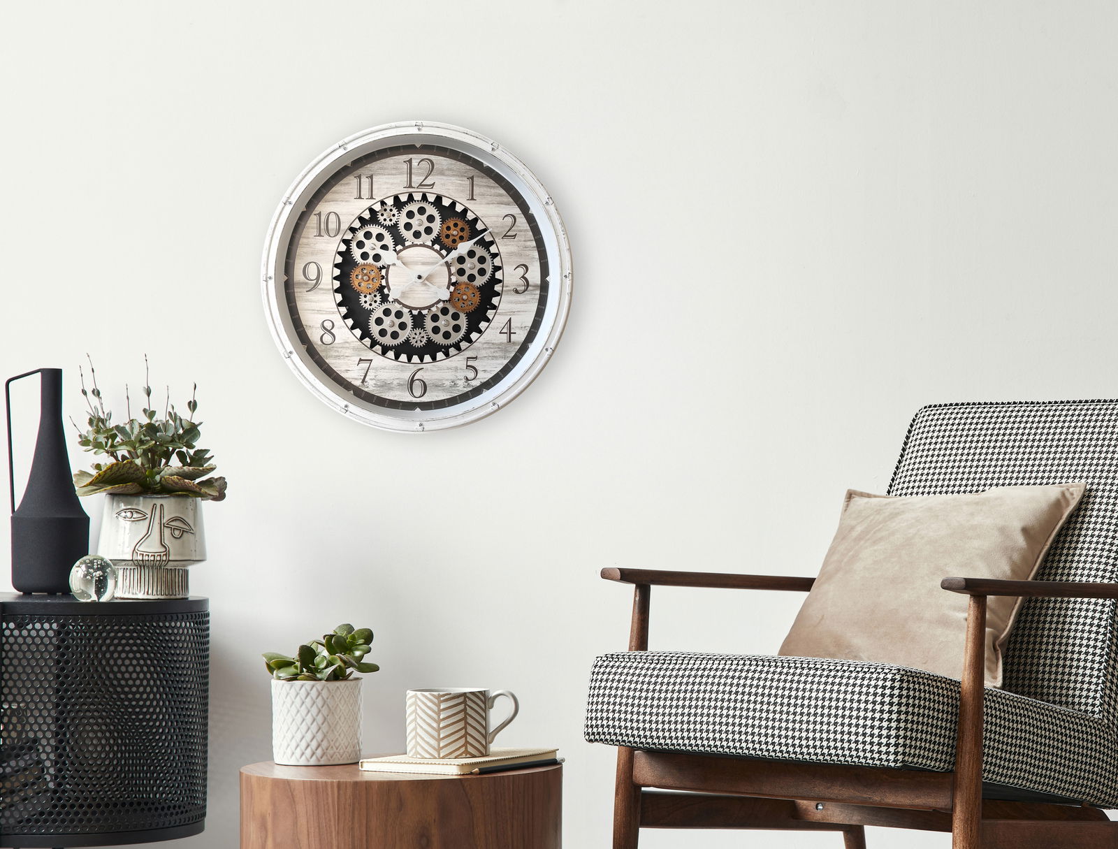 Wanduhr mit Zahnrädern im Vintage-Stil an einer weißen Wand, umgeben von moderner Dekoration und einem Sessel, aus frontaler Perspektive.