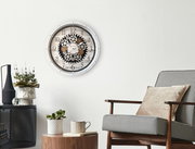 Wanduhr mit Zahnrädern im Vintage-Stil an einer weißen Wand, umgeben von moderner Dekoration und einem Sessel, aus frontaler Perspektive.