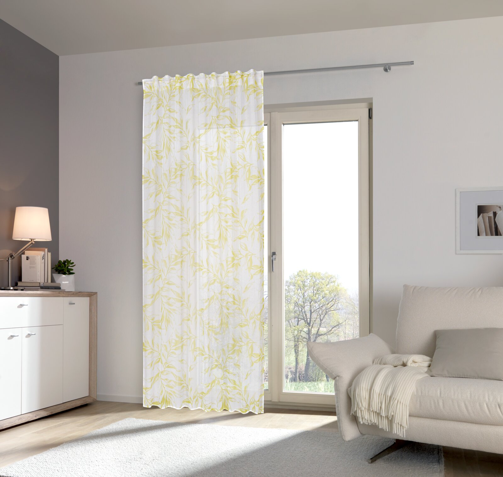 Fertigvorhang Flora 140x245 in einem modernen Wohnzimmer, aufgehängt an einer Gardinenstange vor einer großen Glastür, mit einem hellen, blattförmigen Muster in Gelb und Weiß.