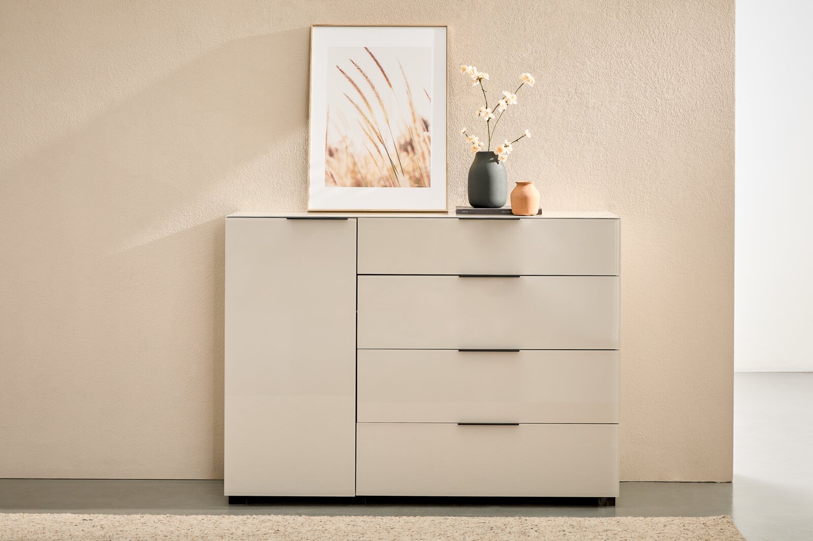 Novel Combinatiecommode MALIN 2.0 Moderne witte ladekast met één deur aan de linkerkant en drie lades aan de rechterkant, voorzien van zwarte handgrepen; daarop een ingelijste poster en twee vazen, van voren gefotografeerd vanuit een licht verhoogd perspectief