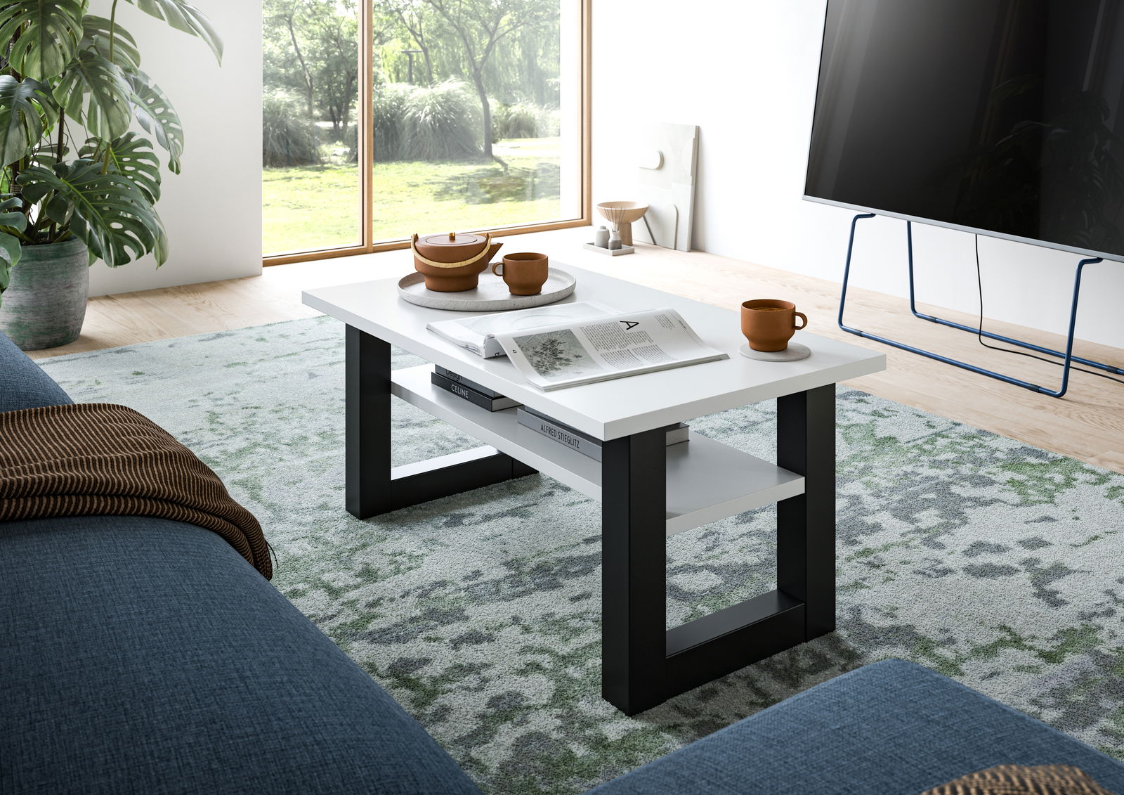 Moderner Couchtisch SEATTLE mit weißer Tischplatte und schwarzen Beinen, seitliche Perspektive im Wohnzimmer mit großen Fenstern und grüner Pflanze.