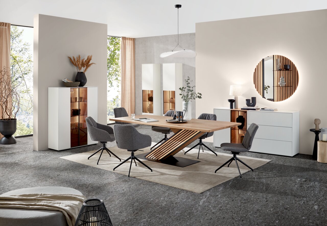 Moderne Esszimmer-Einrichtung mit Esstisch aus Holz, umgeben von grauen Stühlen, und weißen Vitrinen im Hintergrund. Perspektive: Frontalansicht.