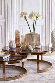 Zwei runde Couchtische mit goldenen Rahmen und Glasplatten, dekoriert mit Vasen und Blumen, aus seitlicher Perspektive fotografiert.