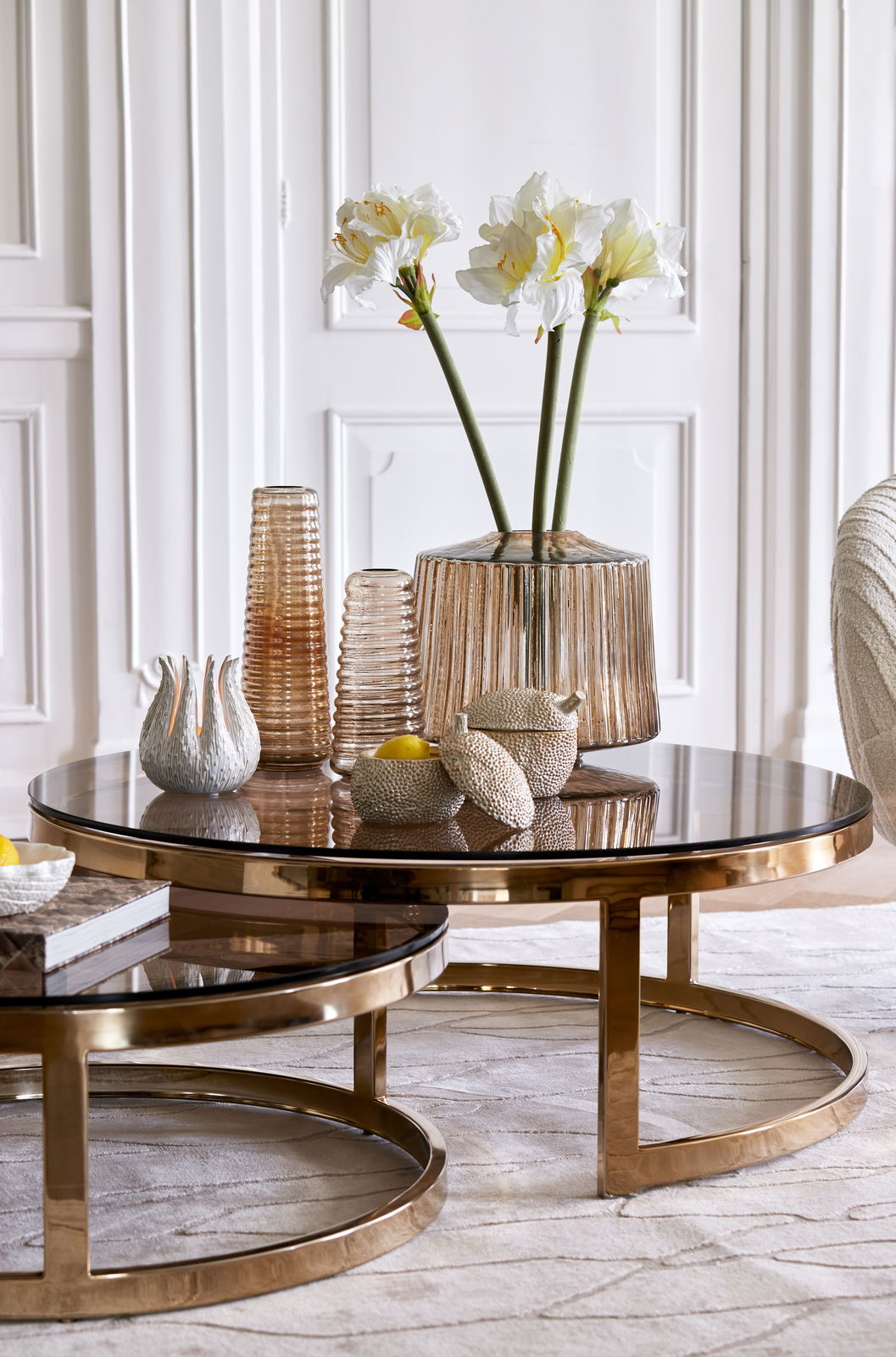 Zwei runde Couchtische mit goldenen Rahmen und Glasplatten, dekoriert mit Vasen und Blumen, aus seitlicher Perspektive fotografiert.