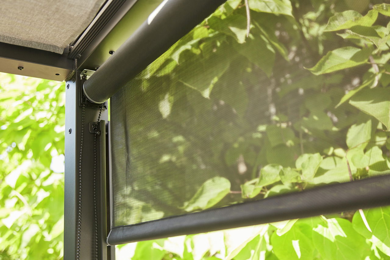 Nahaufnahme eines Rollos mit Kettenzug, montiert an einem Metallrahmen, mit Blick auf grüne Blätter im Hintergrund.