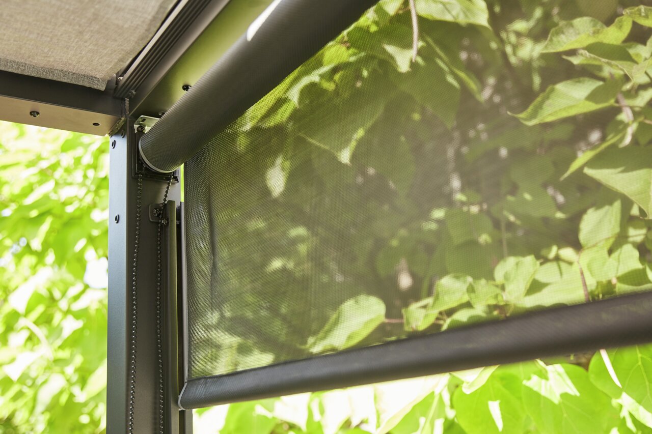 Close-up van een rolgordijn met kettingmechanisme, gemonteerd op een metalen frame, met groene bladeren op de achtergrond.