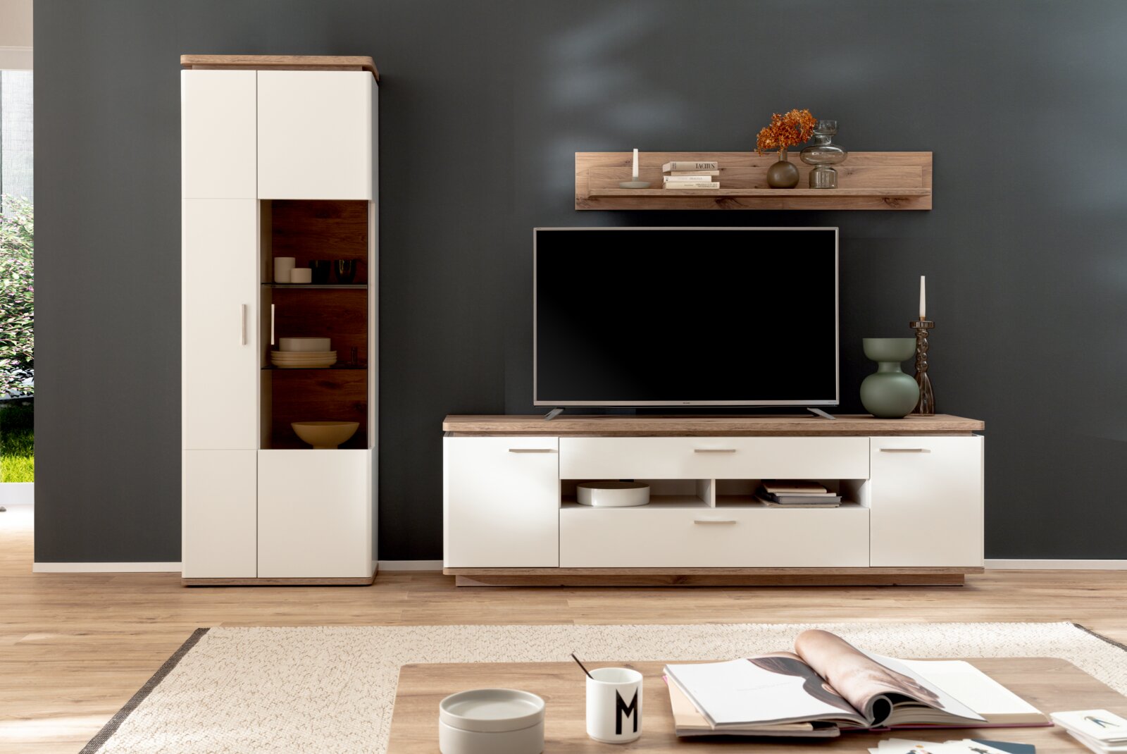 Modernes Wohnzimmer-Entertainment-Set mit weißer TV-Bank und hoher Vitrine mit Holzoptik, Flachbildfernseher und Wandregal auf dunkelgrauer Wand, frontal aus leicht seitlicher Perspektive aufgenommen