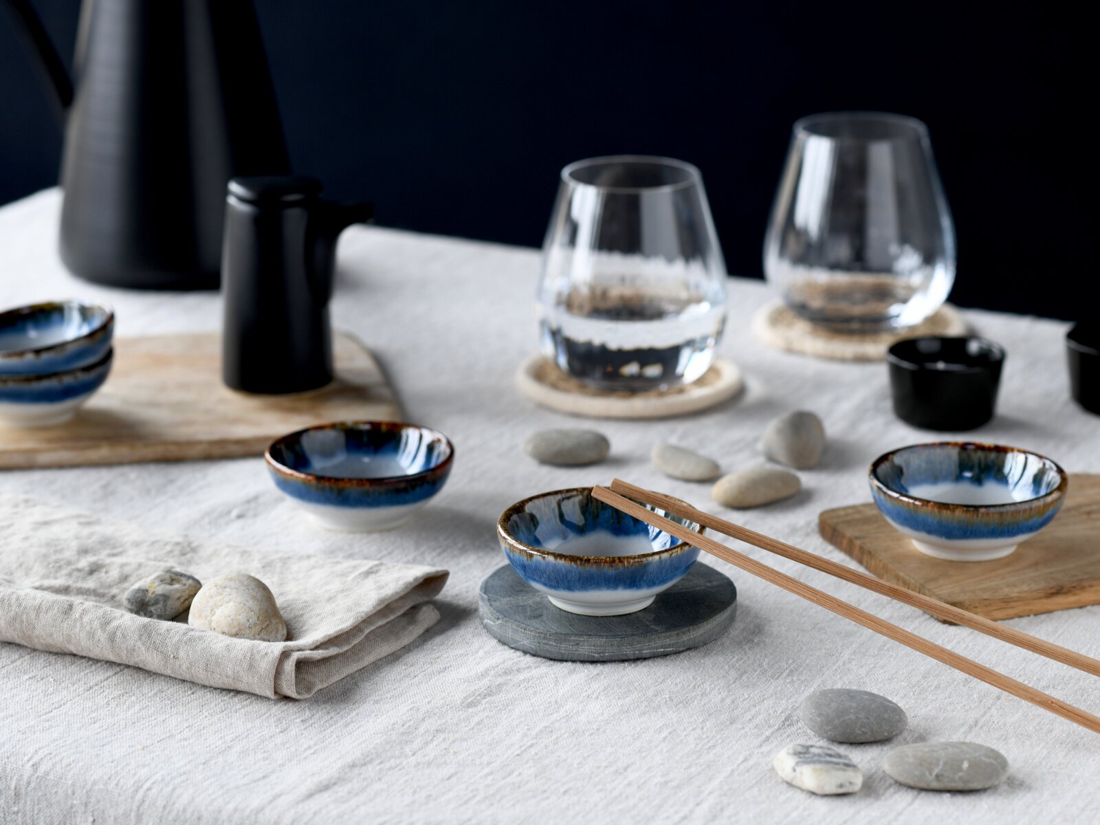 Japanisch inspiriertes Tischarrangement mit kleinen blau glasierten Keramikschälchen, Holzstäbchen und Wassergläsern auf Leinentuch, fotografiert aus leicht erhöhter schräger Perspektive