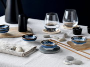 Japanisch inspiriertes Tischarrangement mit kleinen blau glasierten Keramikschälchen, Holzstäbchen und Wassergläsern auf Leinentuch, fotografiert aus leicht erhöhter schräger Perspektive