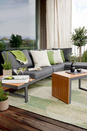 Gartenlounge-Set auf einer Terrasse mit grauem Sofa, grünen und weißen Kissen, einem Holztisch und Pflanzen, seitliche Perspektive.
