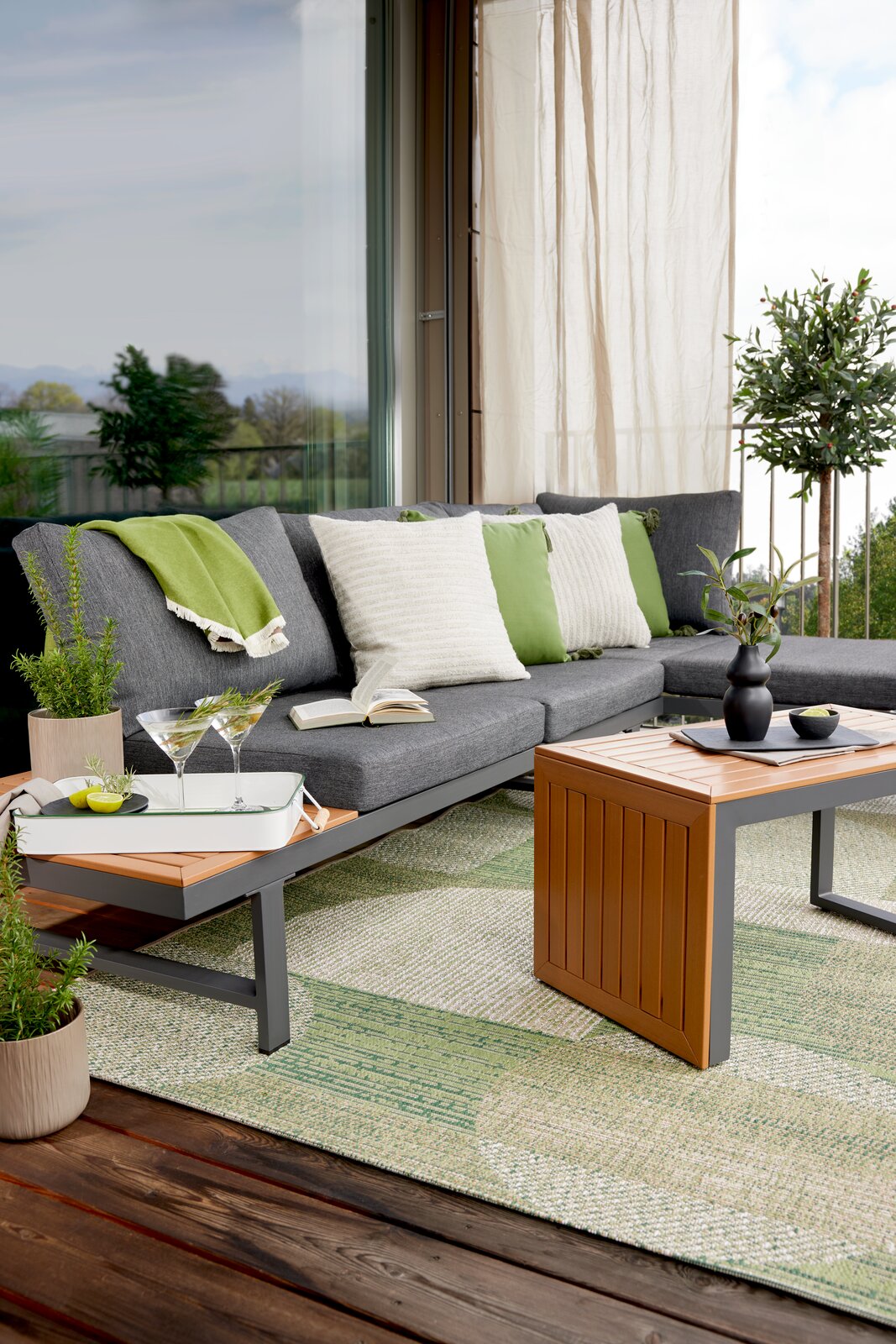 Gartenlounge-Set auf einer Terrasse mit grauem Sofa, grünen und weißen Kissen, einem Holztisch und Pflanzen, seitliche Perspektive.