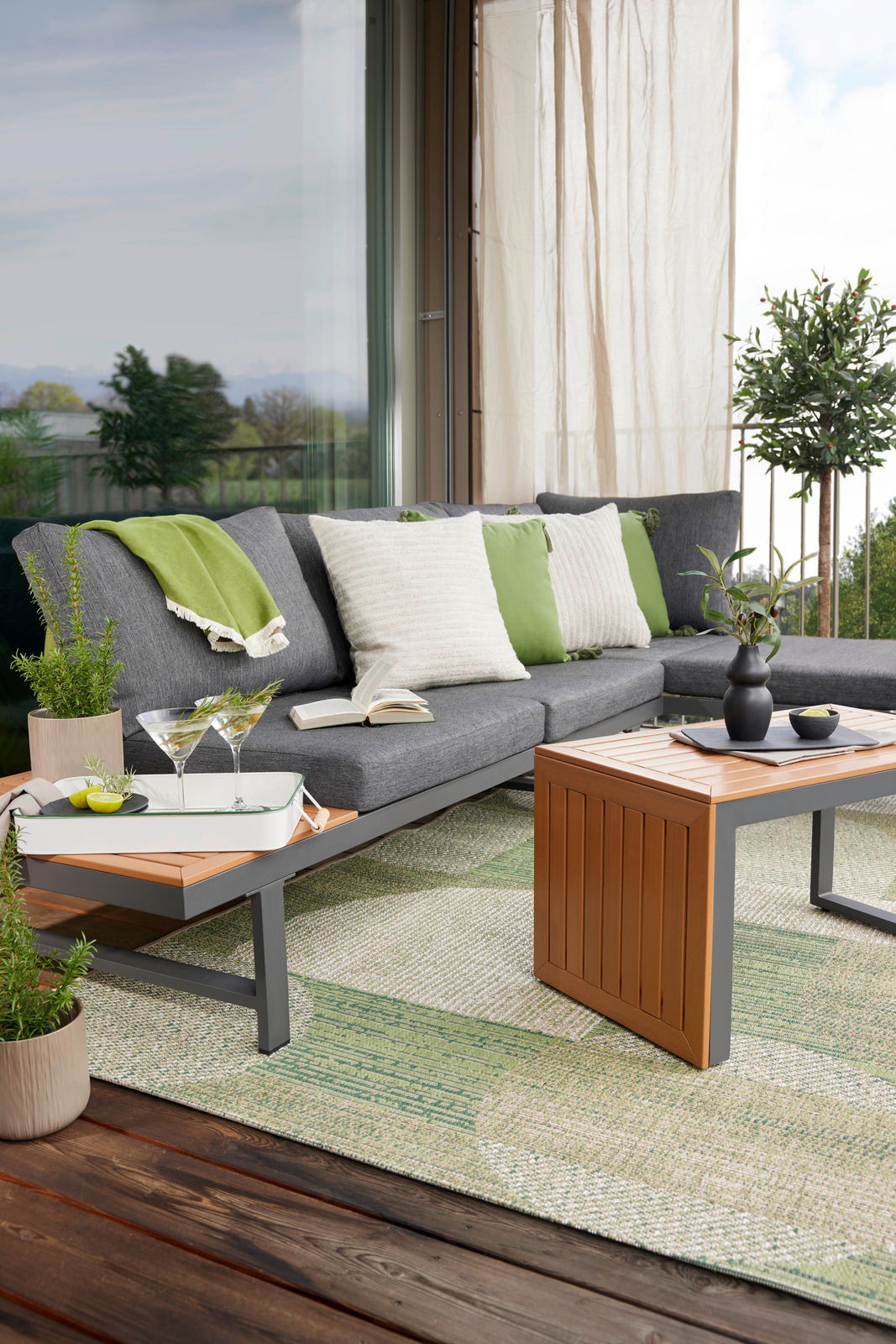 Gartenlounge-Set auf einer Terrasse mit grauem Sofa, grünen und weißen Kissen, einem Holztisch und Pflanzen, seitliche Perspektive.