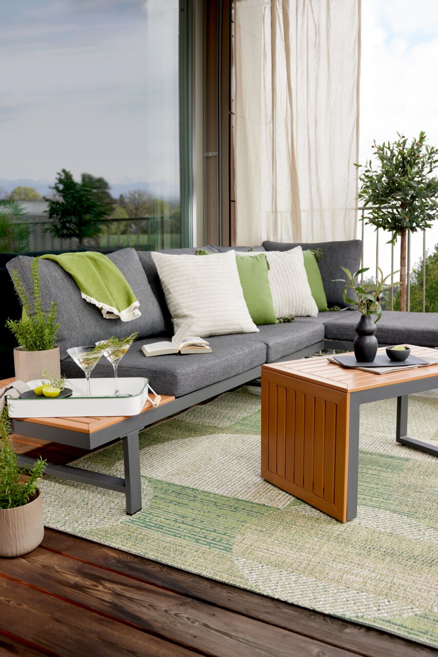 Tuinset op een terras met een grijze bank, groene en witte kussens, een houten tafel en planten, gezien vanaf de zijkant.