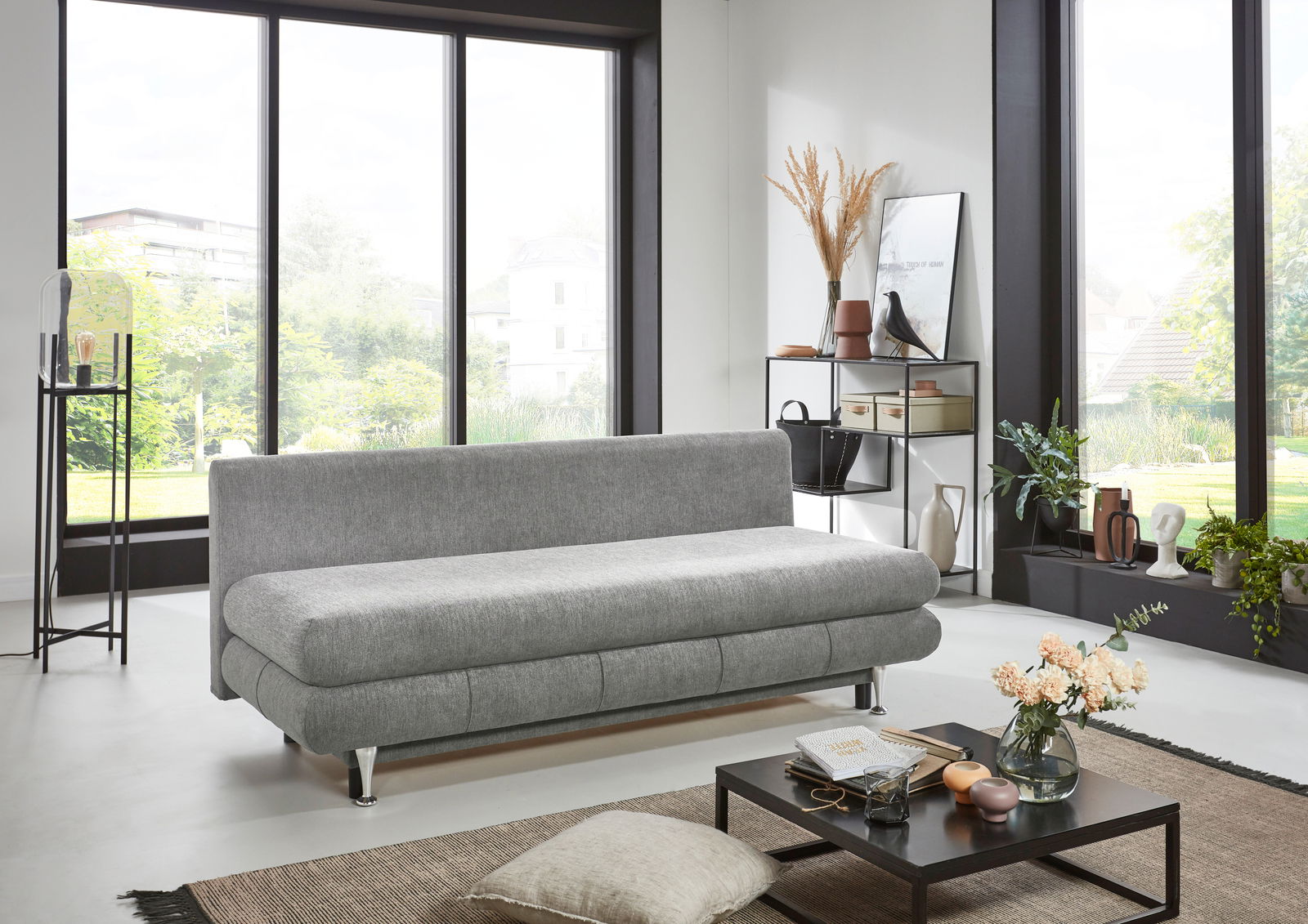 Graues Funktionssofa in modernem Wohnzimmer, seitliche Perspektive mit großen Fenstern im Hintergrund.
