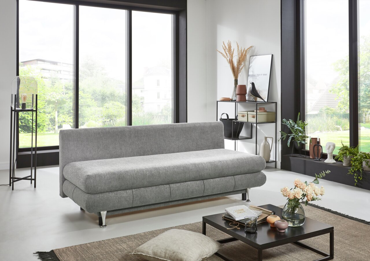 Graues Funktionssofa in modernem Wohnzimmer, seitliche Perspektive mit großen Fenstern im Hintergrund.