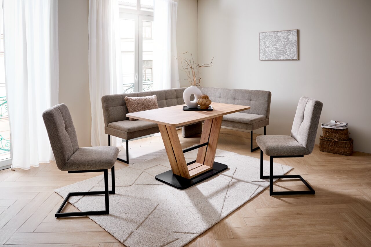 Moderner Esstisch mit V-förmigem Holzgestell, umgeben von grauen Polsterstühlen und einer Eckbank, in einem hellen Raum mit großen Fenstern, aus der Frontalperspektive.