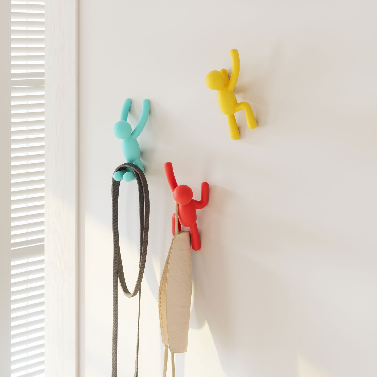 Drei bunte Wandhaken in Form von kletternden Figuren in Blau, Rot und Gelb, die an einer weißen Wand befestigt sind. Perspektive von vorne.