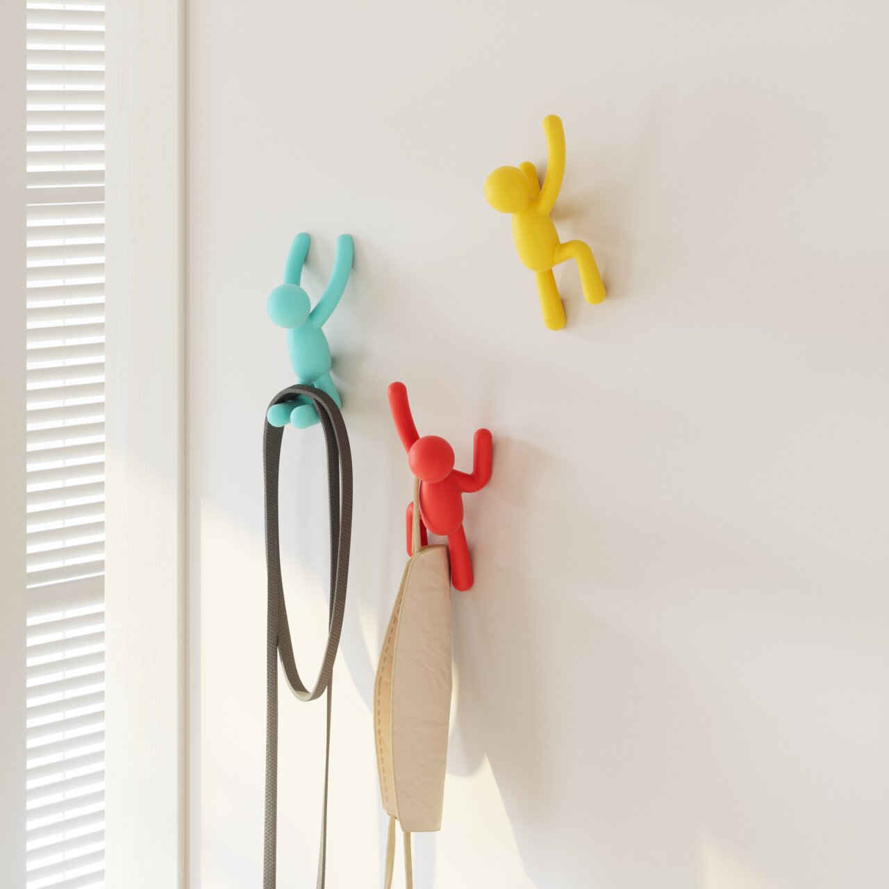 Drei bunte Wandhaken in Form von kletternden Figuren in Blau, Rot und Gelb, die an einer weißen Wand befestigt sind. Perspektive von vorne.