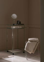 Elegantes Badezimmer mit einem gestapelten Duschtuch auf einem transparenten Stuhl, im Hintergrund ein moderner Waschtisch mit Spiegel und Seifenspender, seitliche Perspektive.