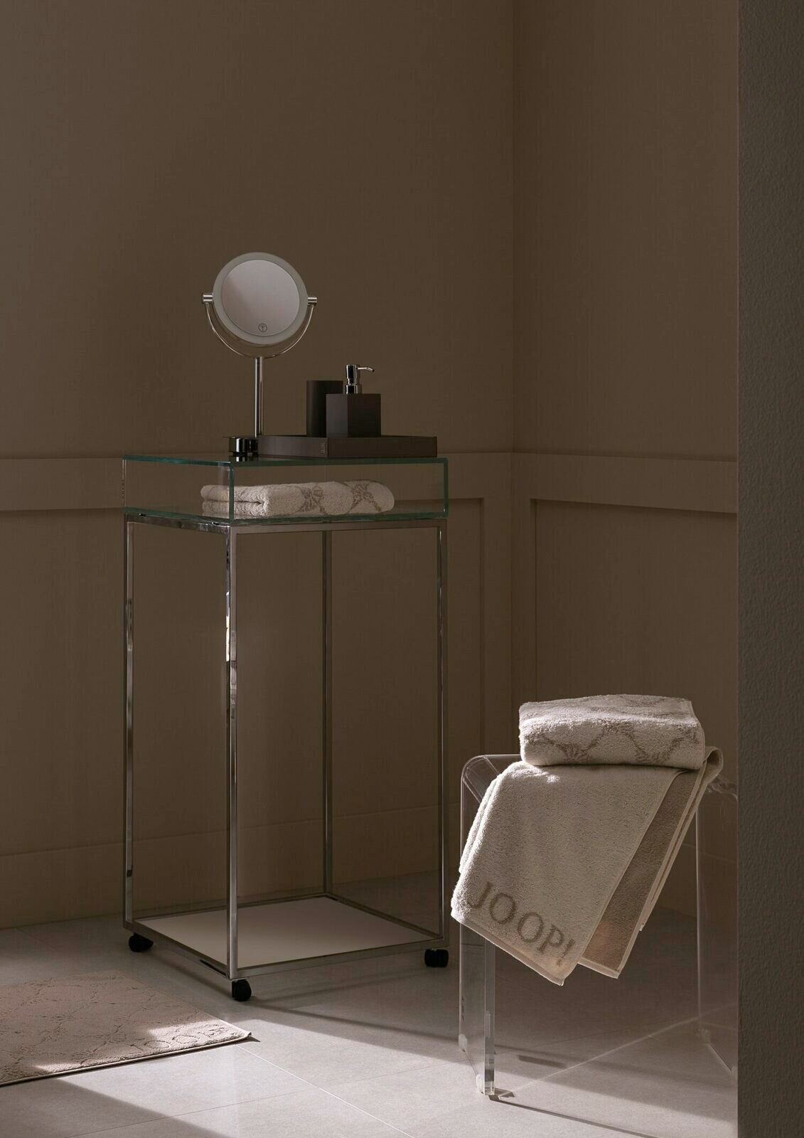 Elegantes Badezimmer mit einem gestapelten Duschtuch auf einem transparenten Stuhl, im Hintergrund ein moderner Waschtisch mit Spiegel und Seifenspender, seitliche Perspektive.