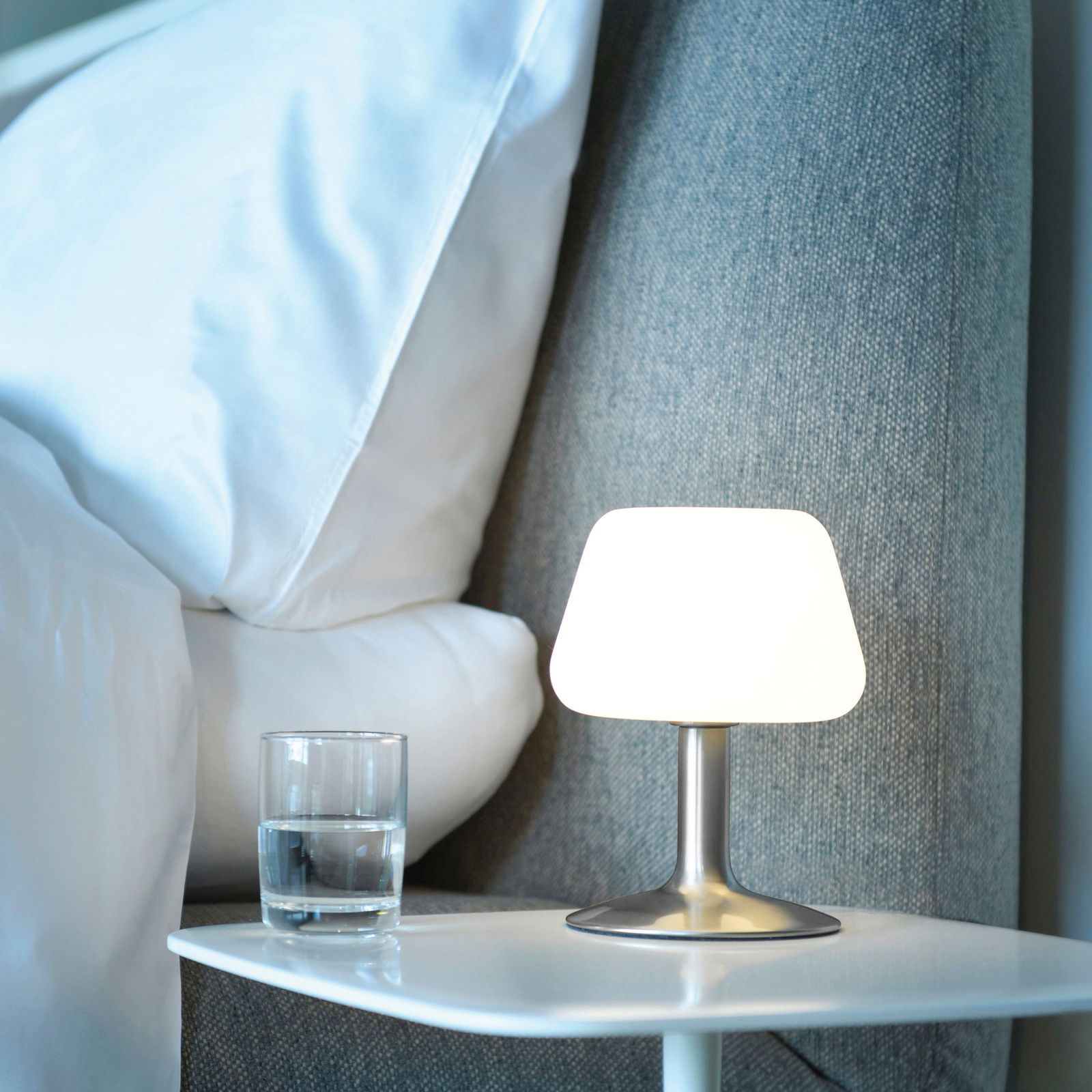 Moderne Tischleuchte aus Stahl auf einem Nachttisch, seitliche Perspektive, neben einem Glas Wasser und einem Bett.