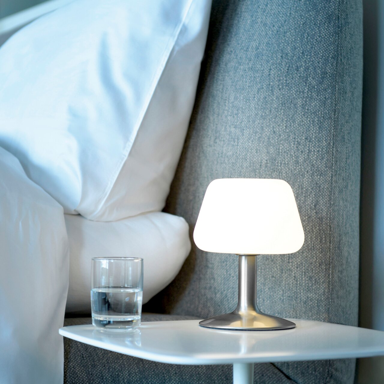 Moderne Tischleuchte aus Stahl auf einem Nachttisch, seitliche Perspektive, neben einem Glas Wasser und einem Bett.
