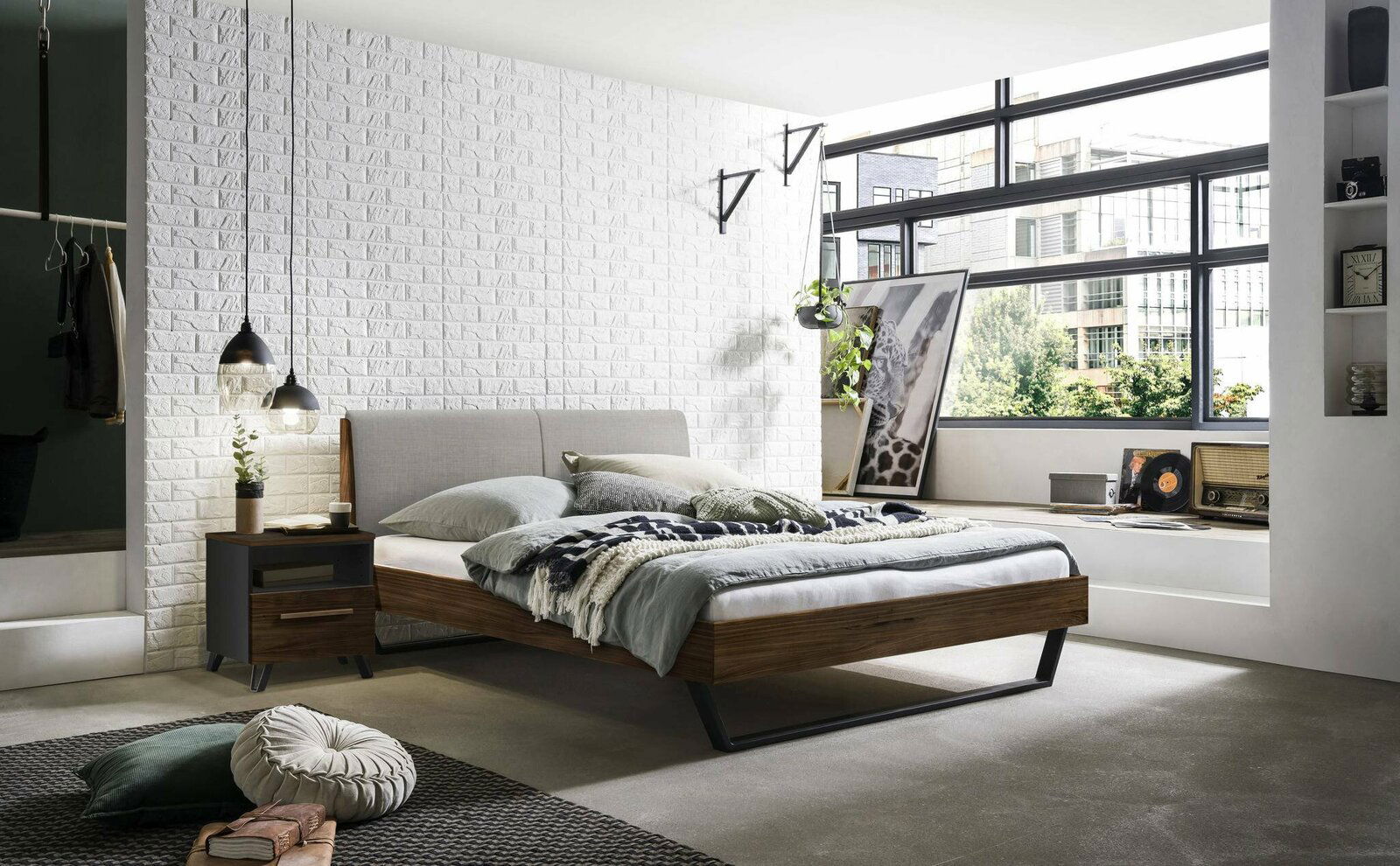 Modernes Schlafzimmer mit einem stilvollen Bettgestell aus Holz und Metall, seitliche Perspektive. Das Bett ist mit grauer Bettwäsche und dekorativen Kissen ausgestattet, umgeben von einer weißen Ziegelwand und großen Fenstern, die viel Tageslicht hereinl