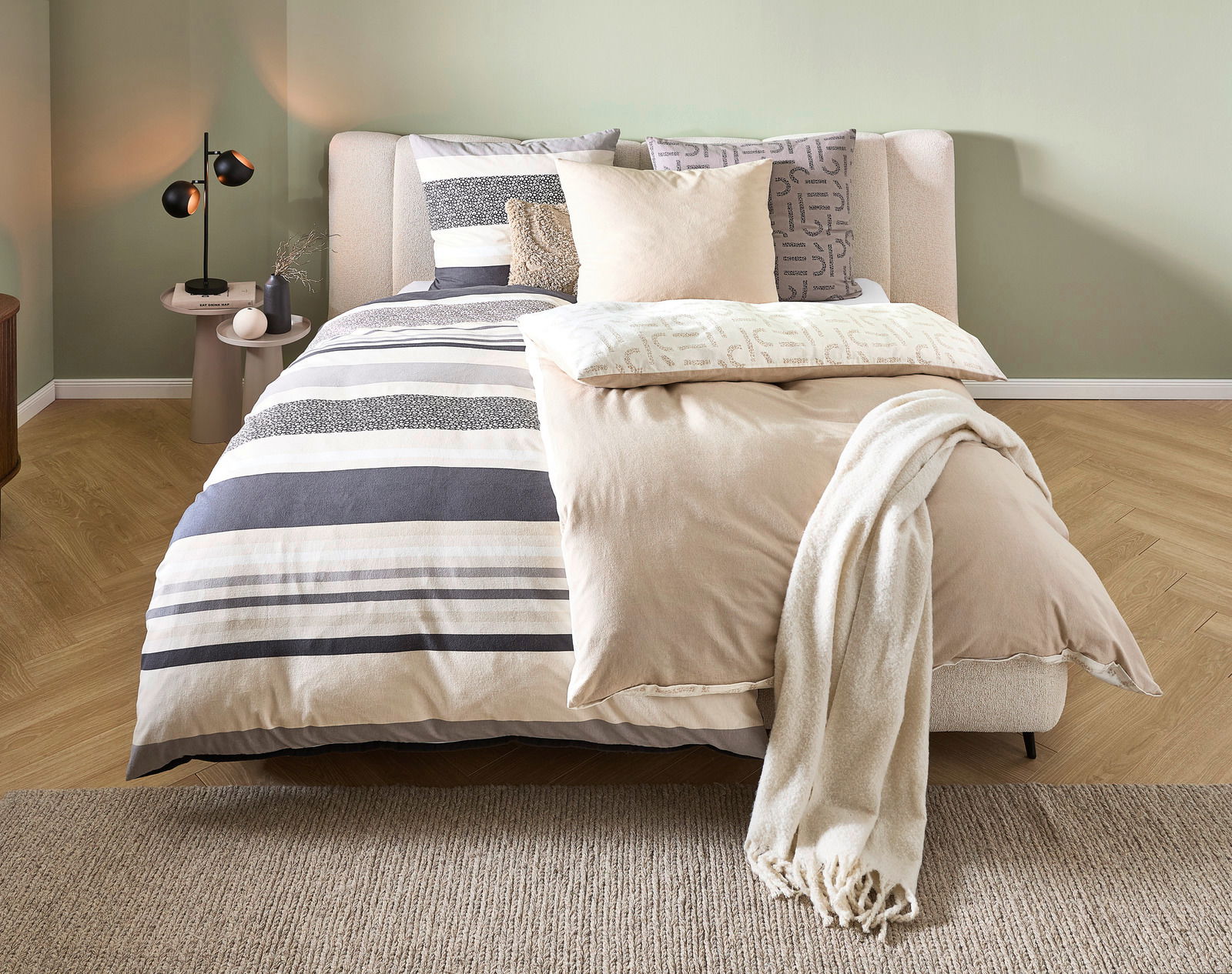 Gemütliches Schlafzimmer mit modernem Bett, das mit gestreifter Bettwäsche in Beige- und Grautönen bezogen ist. Perspektive von vorne.