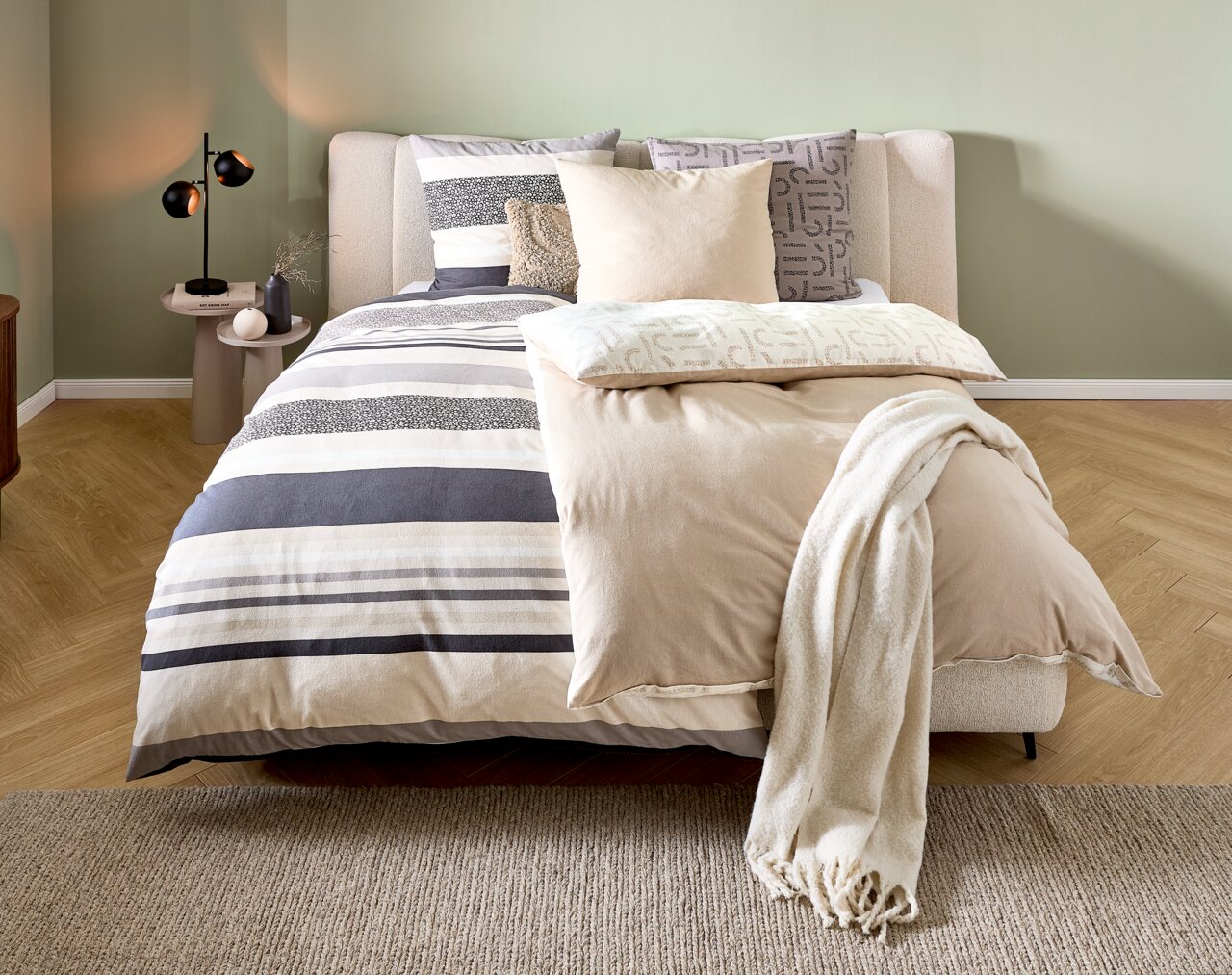 Gemütliches Schlafzimmer mit modernem Bett, das mit gestreifter Bettwäsche in Beige- und Grautönen bezogen ist. Perspektive von vorne.