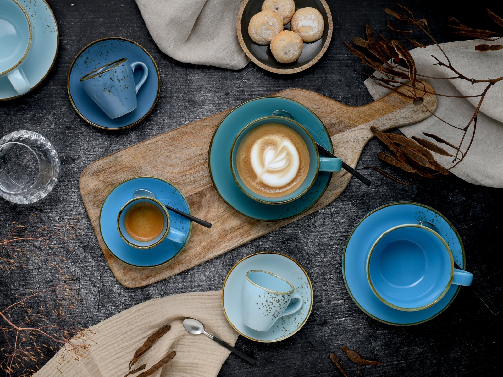 Draufsicht auf ein blaues Keramik-Kaffeeservice mit Tassen und Untertassen auf rustikalem Holztisch, Cappuccino mit Latte Art auf Holzbrett, dazu Gebäck und Leinentuch.