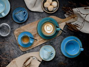 Draufsicht auf ein blaues Keramik-Kaffeeservice mit Tassen und Untertassen auf rustikalem Holztisch, Cappuccino mit Latte Art auf Holzbrett, dazu Gebäck und Leinentuch.