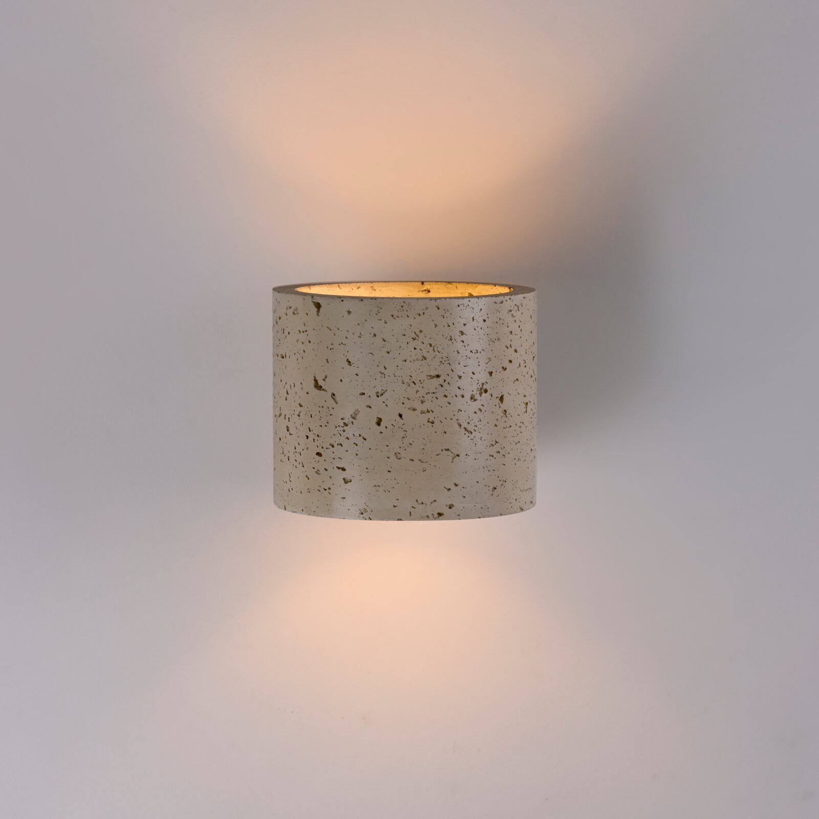 Zylindrische Wandlampe aus gesprenkeltem Beton in Hellgrau mit warmem indirektem Lichtschein nach oben und unten, frontal in leichter Untersicht vor neutralem Hintergrund fotografiert