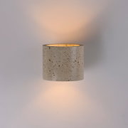 Zylindrische Wandlampe aus gesprenkeltem Beton in Hellgrau mit warmem indirektem Lichtschein nach oben und unten, frontal in leichter Untersicht vor neutralem Hintergrund fotografiert