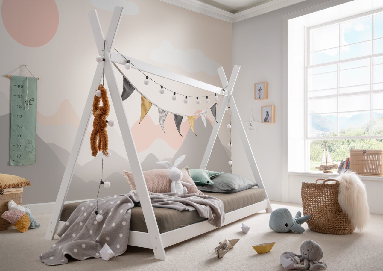 Tipibett aus weißem Holz mit dekorativen Lichterketten und Wimpeln, umgeben von Kuscheltieren und Spielzeug in einem hellen Kinderzimmer mit großem Fenster.