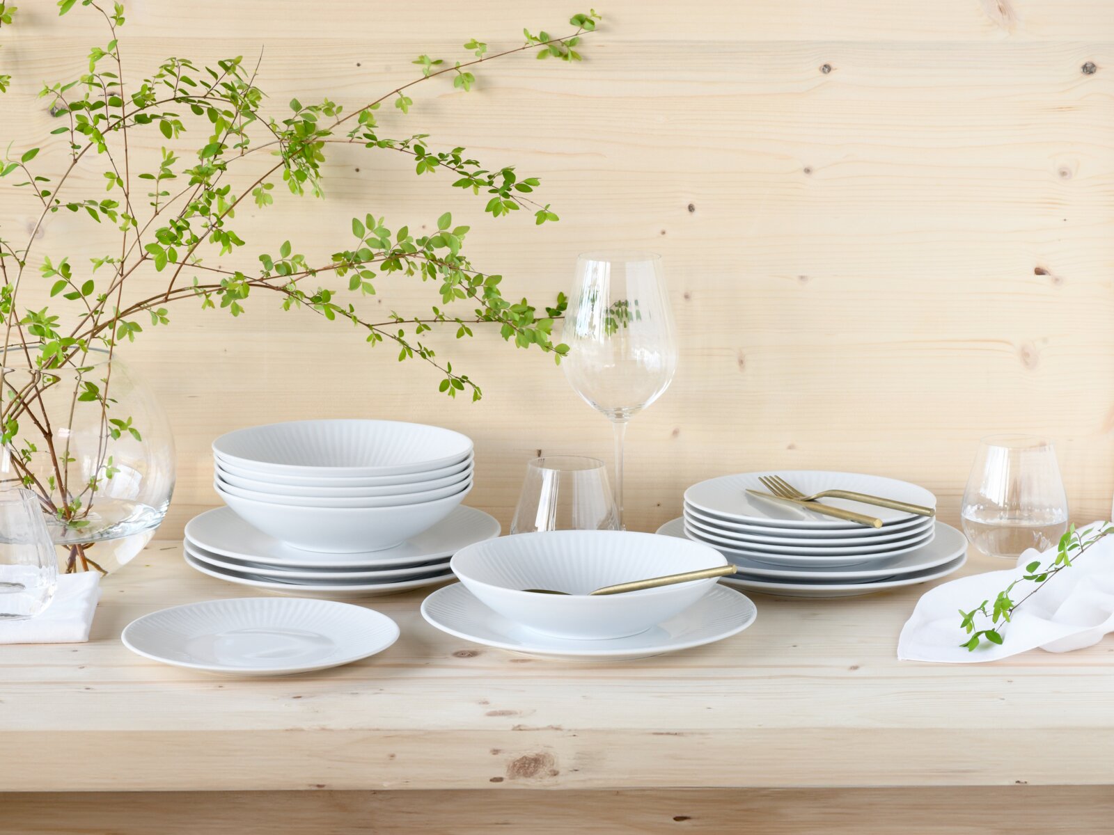 Creatable Tellerset 18-tlg VALENCIA Weißes, geriffeltes Porzellan-Geschirrset mit gestapelten Tellern und tiefen Schalen auf hellem Holztisch, ergänzt durch Glasvasen, Weingläser und goldfarbenes Besteck, frontal in leicht erhöhter Perspektive.