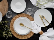 Draufsicht auf ein elegant gedecktes Tisch-Setting mit weißen, geriffelten Porzellantellern und -schalen, goldfarbenem Besteck, geflochtenem Platzset, Glasuntersetzern und weißen Servietten auf dunkler Tischplatte