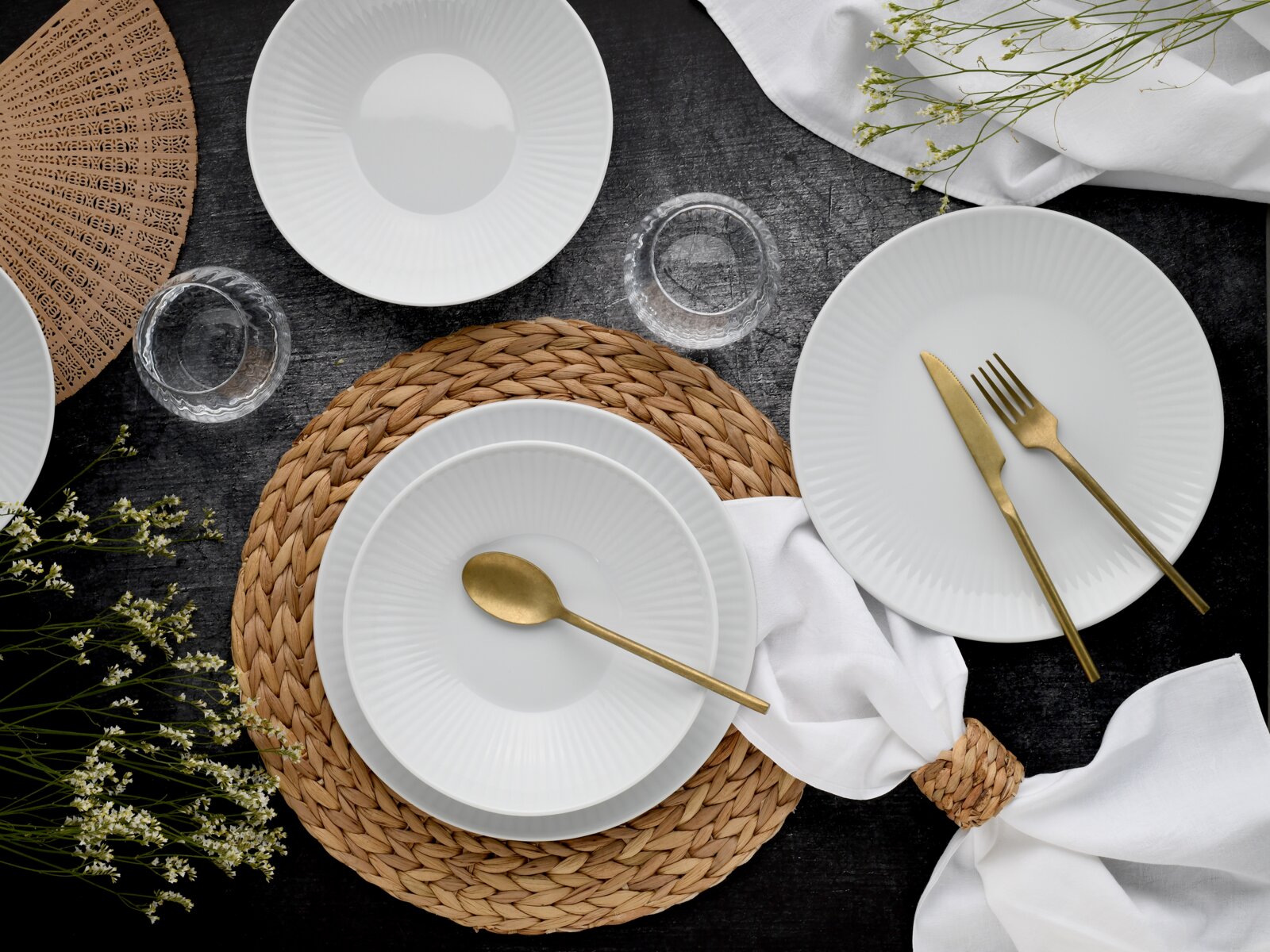 Draufsicht auf ein elegant gedecktes Tisch-Setting mit weißen, geriffelten Porzellantellern und -schalen, goldfarbenem Besteck, geflochtenem Platzset, Glasuntersetzern und weißen Servietten auf dunkler Tischplatte