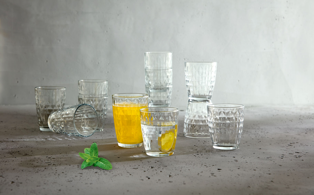 Ein 12-teiliges Gläserset mit geometrischem Muster, teilweise gefüllt mit Getränken, auf einer grauen Oberfläche. Perspektive von vorne.