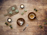 Draufsicht auf rustikalem Holztisch: zwei Espressotassen auf Untertassen, ein Löffel, zwei brennende Teelichter, grüne Kekse auf Holzbrett (einer angebissen) und mehrere verpackte Espresso-Bohnen-Pralinen.