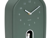Grüne Wanduhr mit weißen Ziffern und Zeigern, zeigt die Zeit 2:10 an, mit einer kleinen weißen Katze in einer Öffnung unten. Perspektive von schräg vorne.